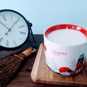 Op de afbeelding: Een witte kaars in een mok met een rode binnenkant en de woorden "Kansas City" op de zijkant gedrukt. Een zwarte klok en een kleine bezem staan ook op het houten oppervlak.