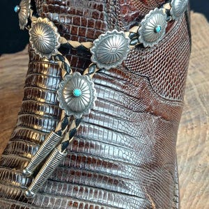 May include: Close-up of a brown leather cowboy boot with intricate stitching and a textured pattern. A silver bolo tie with turquoise accents and decorative silver conchos is draped over the boot, showcasing Western style.