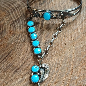 May include: A silver slave bracelet featuring a cuff with a central turquoise stone and leaf accents, connected by a chain of turquoise stones to a ring. The ring also features two turquoise stones and a silver leaf design, with a matching chain.