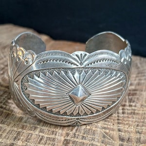 May include: A wide silver cuff bracelet featuring an intricate stamped design. The center displays a raised diamond shape surrounded by radiating sunburst lines on a textured background. The edges are scalloped with detailed engravings.