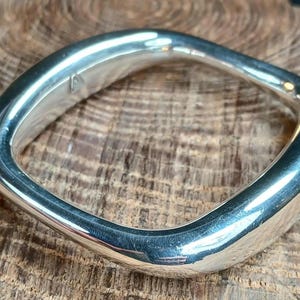 May include: A polished silver bangle with a rounded square shape and a smooth, reflective surface. The metal band features a subtle, irregular contour along its edges, creating a soft, organic form that catches the light across its metallic finish.