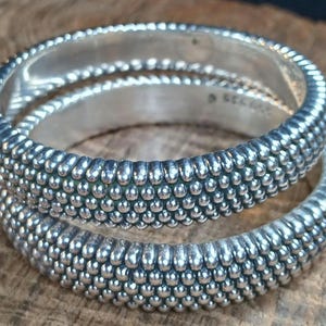 May include: Two silver rings with a textured, beaded design. The rings are stacked, showcasing the intricate detail of the small, rounded beads that cover the bands. The rings are on a wooden surface.