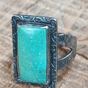 May include: A silver ring with a rectangular turquoise stone. The stone is set in a decorative frame with an ornate pattern. The ring's band has a textured design, and the ring is displayed on a wooden surface.