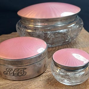 May include: Three antique vanity jars. The jars have silver rims and pink enamel lids with a textured design. Two jars have clear glass bodies with floral etchings, and one is a silver box with engraved initials. The jars are on a wooden surface.