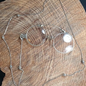 May include: A pair of silver-toned pince-nez spectacles featuring round clear lenses and a delicate metal bridge. The eyewear is attached to a long, thin silver-toned chain that includes small, decorative spherical beads spaced along its length.