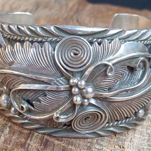 May include: A silver cuff bracelet with intricate floral and spiral designs. The detailed metalwork features leaves, berries, and coiled elements. The open cuff design allows for easy wear. A beautiful piece of jewelry.