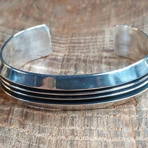 May include: A silver cuff bracelet with three black horizontal lines. The bracelet has a polished finish and a simple, modern design. The open ends of the bracelet are rectangular.