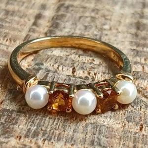 May include: A gold ring featuring an alternating arrangement of three round white pearls and two faceted orange-brown gemstones. The band is polished gold, and the stones are set in a row across the top section of the circular metal band.
