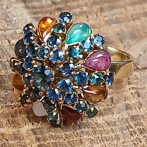 May include: A gold-toned ring featuring a cluster of colorful gemstones. The ring showcases a variety of stones in shades of blue, green, orange, and purple. The gemstones are arranged in a floral or cluster design, creating a bold and eye-catching piece of jewelry.