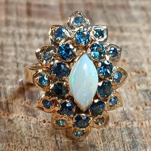 May include: A gold ring featuring a central, pear-shaped opal surrounded by a cluster of blue gemstones. The ring's setting has a floral design, with the blue stones arranged around the opal. The ring is set on a wooden surface.