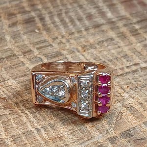 May include: Rose gold ring with a rectangular design. The ring features a pear-shaped setting with clear stones, a rectangular section with clear stones, and a section with three red stones. The ring is set on a wooden surface.