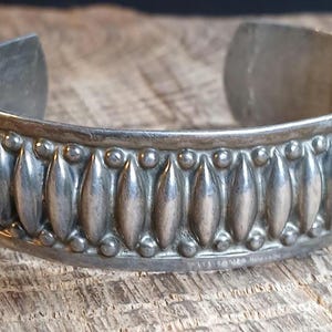 May include: A silver-toned cuff bracelet featuring a repeating pattern of vertical, elongated oval shapes bordered by small, raised spherical beads along the top and bottom edges. The metal has a polished finish with visible oxidation in the crevices.