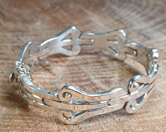 May include: A silver ring with an intricate design featuring repeating curved and geometric shapes. The ring is made of a shiny metal and appears to be handcrafted. The ring is displayed on a wooden surface.