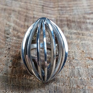 May include: A silver-colored ring with a cage-like design. The ring features vertical bars that curve to form an oval shape. The ring is set against a wooden background.