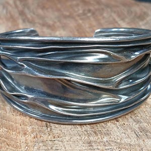 May include: A silver bracelet with a wavy, organic design. The bracelet is made of multiple layers of metal that have been hammered and shaped to create a flowing, textured surface.