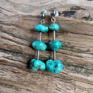 May include: Turquoise dangle earrings with three turquoise beads on each earring. The earrings are silver and have a silver hook closure.
