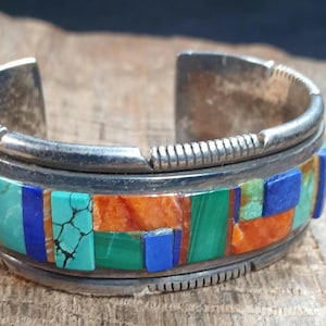 May include: A silver cuff bracelet with a mosaic design. The mosaic features turquoise, blue, green, and orange rectangular stones. The bracelet has a textured edge and a vintage aesthetic.