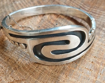 May include: A silver-toned hinged bangle bracelet with a geometric design. The bracelet features a central, stylized swirl pattern and is secured with a small chain. The metal has a polished finish, and the bracelet is displayed on a wooden surface.