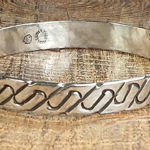 May include: A silver-toned metal bangle bracelet featuring a continuous embossed wave or link pattern around the exterior. The interior surface is smooth and includes a small stamped hallmark with the text MEXICO and a circular emblem.