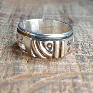 May include: A silver spinner ring featuring a wide band with a polished finish. The exterior surface displays a central section with alternating vertical and diagonal gold-toned metal bars, accented by small circular cutouts along the edges.