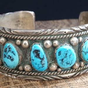 May include: A silver cuff bracelet featuring a series of oval-shaped turquoise stones with dark matrix patterns. The metal band is decorated with twisted wire borders, small silver spheres, and engraved leaf-like details along the edges.