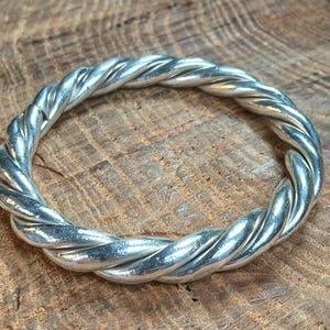 May include: A silver-colored ring with a braided rope design. The ring is circular and appears to be made of metal. The ring is displayed on a wooden surface.