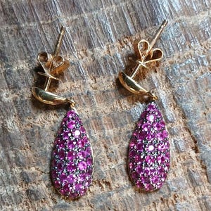 May include: A pair of gold stud earrings featuring teardrop-shaped pendants encrusted with numerous small, vibrant pink stones. Each earring includes a gold post backing and a curved gold element connecting the stud to the teardrop drop.