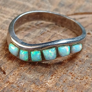 May include: A silver ring with six rectangular opal stones. The opals have a play of color, showing flashes of green and blue. The ring is set on a textured, brown surface.
