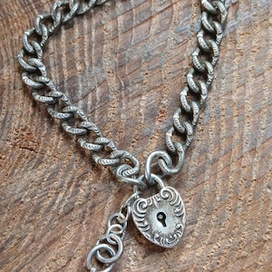 May include: A silver-toned metal chain bracelet featuring a heart-shaped padlock charm with intricate embossed scrollwork and a central keyhole. A small, slender metal key is attached to the padlock by a series of three small circular jump rings.
