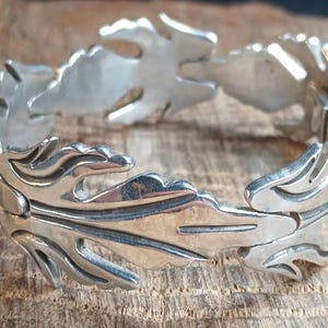 May include: A silver-toned link bracelet composed of repeating leaf-shaped segments. Each individual link features a polished surface with recessed lines that define the organic, pointed shape of the leaves. The bracelet includes a secure clasp.