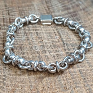 May include: A silver-toned chain bracelet composed of repeating circular links connected by smaller, smooth cylindrical spacers. The bracelet features a rectangular box-style clasp mechanism that secures the circular chain links together.