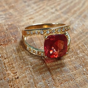 May include: A gold ring featuring a large, square-cut, red gemstone. The band is split, with one side encrusted with small, clear gemstones. The ring is set against a textured, brown surface.