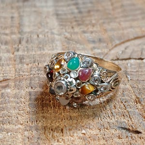 May include: A silver-toned ring with a cluster of colorful gemstones. The ring features a variety of stones in shades of red, green, yellow, and clear, set in a detailed, ornate design. The band has a gold tone.