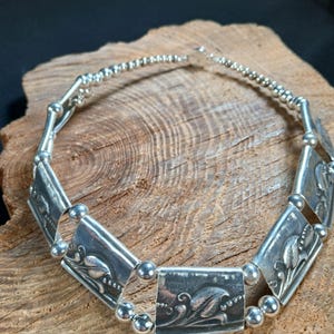 May include: A silver-toned necklace featuring rectangular and diamond-shaped links with floral designs. The necklace is connected by small, round beads and is displayed on a wooden surface.