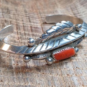May include: A silver cuff bracelet featuring a polished finish with engraved leaf patterns, small spherical silver accents, and a single rectangular orange coral stone set in a serrated bezel. The metal band is open at the back for adjustment.