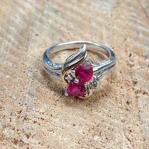 May include: A silver ring with a gold accent features two heart-shaped, deep red gemstones. The ring has a split shank design and small accent stones near the main gemstones. The ring is set on a textured surface.