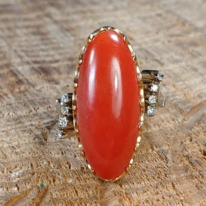 May include: A gold ring featuring a large, oval-shaped, polished red coral stone. The coral is set in a gold bezel, and the ring has additional small, clear stones on the sides. The ring is displayed on a wooden surface.