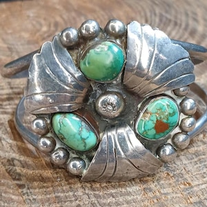 May include: A silver cuff bracelet featuring a circular centerpiece with three oval turquoise stones. The metalwork includes embossed leaf-like patterns and small silver spheres surrounding the stones. A small silver bead with a stamped flower design sits in the center of the arrangement.
