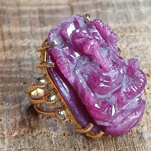 May include: A gold ring with a carved pink ruby gemstone depicting a deity. The ruby is set in a gold frame and the ring has a decorative band.