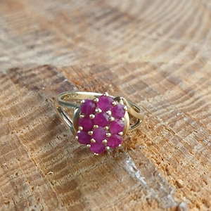 May include: A gold ring featuring a cluster of vibrant, round, ruby gemstones. The ring's band has a split design, and the gemstones are set in a circular arrangement, creating a floral appearance. The ring is displayed on a textured wooden surface.