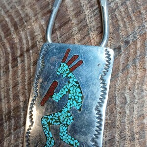 May include: A rectangular silver pendant featuring a Kokopelli figure inlaid with turquoise and red stone fragments. The metal surface has a textured, zigzag-patterned border and a smooth silver bail at the top for attachment to a chain or cord.