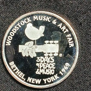 May include: A silver-colored commemorative coin for the Woodstock Music & Art Fair. The coin features a dove on a guitar, the text "3 Days of Peace & Music", and the location "Bethel, New York 1969".