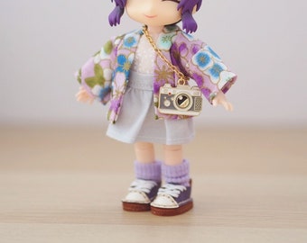 Op de afbeelding: Een kleine pop die een paars-witte bloemenkimono draagt met een camerabedel. De pop heeft paars haar en draagt witte sokken en paarse sneakers.