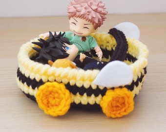 Op de afbeelding: Twee kleine poppen, een met roze haar en een met zwart haar, zitten in een gehaakte bij-vormige mand. De mand is geel en zwart met witte vleugels en heeft gele bloemenaccenten.