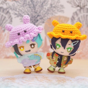May include: Two crocheted plush toys with smiling faces. The toy on the left is wearing a purple and pink crocheted hat and a light blue shirt with a fish design. The toy on the right is wearing an orange crocheted hat and a green and gold patterned shirt.