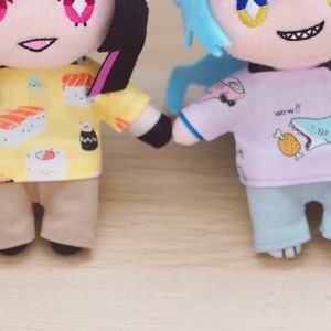 Könnte beinhalten: Zwei Plüschtiere, eines mit schwarzen Haaren und einem gelben Shirt mit Sushi-Motiven, das andere mit blauen Haaren und einem rosa Shirt mit einem Hai-Motiv. Beide Spielzeuge tragen Hosen.