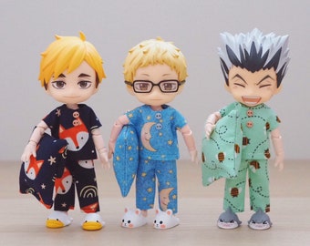 Op de afbeelding: Drie kleine poppen die pyjama's dragen. De pop links draagt een blauw en oranje pyjama met een vos print. De pop in het midden draagt een blauwe pyjama met een maan en sterren print. De pop rechts draagt een groene pyjama met een bij print.