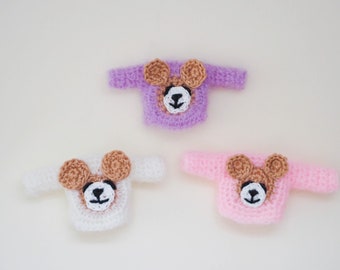 May include: Three crocheted sweaters for dolls, each featuring a brown bear face with black eyes and a black nose. The sweaters are in purple, white, and pink.