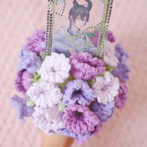 Puede incluir: Un ramo de flores de ganchillo en tonos morados y lavanda, con una imagen enmarcada de un personaje de anime. El marco está adornado con pedrería. El ramo se sostiene en una mano.