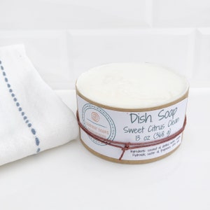 Op de afbeelding: Een ronde afwaszeep met een Sweet Citrus Clean geur, verpakt in een kartonnen container. Het etiket bevat de tekst "Dish Soap" en "368 g". Links ligt een witte doek met blauwe stiksels.